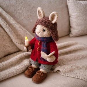 May include: A handmade, knitted bunny doll wearing a red coat, brown hat, and blue scarf. The bunny holds a candle and wears brown shoes and gray pants. The doll is sitting on a beige blanket.