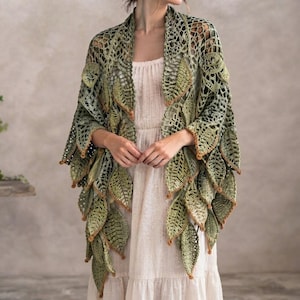 May include: A crochet shawl featuring a complex openwork pattern with numerous dangling three-dimensional leaf motifs in various shades of green and brown. The garment is draped over a simple, sleeveless, cream-colored dress with a tiered skirt.
