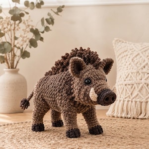 May include: A brown crocheted wild boar figurine featuring a textured mane, dark brown hooves, a dark snout, and two small cream-colored tusks. The stuffed animal has black plastic eyes and a small tail with a tuft of yarn at the end.