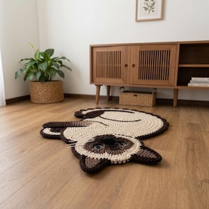 May include: A crochet rug shaped like a Siamese cat with a cream-colored body, dark brown ears, face, and tail. The cat has blue eyes, a black nose, and white whiskers. The rug is crafted with thick yarn in a circular pattern, lying flat on the floor.