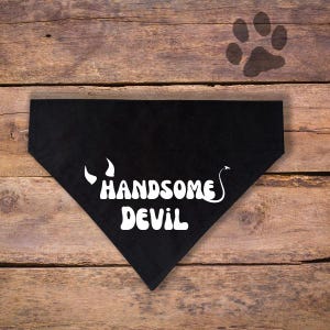 May include: Black triangular pet bandana with the words "HANDSOME DEVIL" in white, along with devil horn and tail graphics. The bandana is on a wooden surface with a paw print.