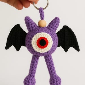 May include: A handmade crochet keychain featuring a purple monster with a large eye, black wings, and small legs. The eye has a white, red, and black design. A wooden bead and metal ring are attached for use as a keychain.