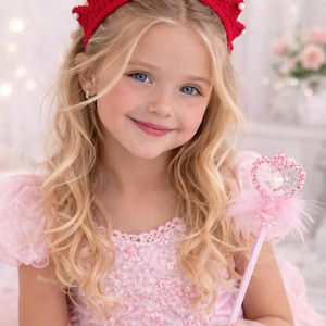 Könnte beinhalten: Ein junges Mädchen mit blonden Haaren und blauen Augen trägt eine rote Krone mit weißen Perlenakzenten. Sie trägt ein rosa Kleid und hält einen rosa Feenstab mit herzförmiger Spitze.