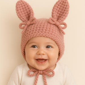 Puede incluir: Un bebé con un gorro de conejito rosa tejido con orejas largas y lazos. El gorro se sujeta con una cinta debajo de la barbilla. El bebé sonríe y lleva un body blanco.