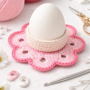 May include: A white egg sits in a cream-colored crocheted egg cup, placed on a pink and white crocheted flower-shaped coaster. Pink yarn balls and crafting tools are in the background. The scene is set on a white surface.