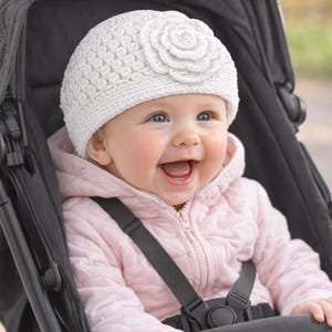Peut inclure: Un bébé souriant dans une poussette noire, portant un bonnet blanc en tricot avec une fleur et une veste rose. Le bébé a les yeux bleus et sourit. La poussette a des sangles noires.