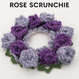 Könnte beinhalten: Ein handgefertigtes Scrunchie mit dem Text "ROSE SCRUNCHIE" oben. Das Scrunchie ist mit gehäkelten lila und grauen Rosen und grünen Blättern verziert. Das Scrunchie ist ein Haaraccessoire.