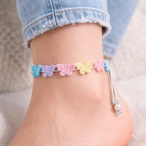 May include: A colorful crochet butterfly anklet. The anklet features six pastel-colored butterflies in blue, purple, pink, yellow, and light blue. A silver-colored string with a pearl and blue bead dangles from the anklet.