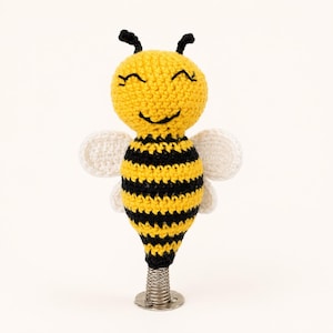 May include: A crocheted bee toy featuring a round yellow head with embroidered black eyes and a smile, a striped yellow and black body, and two white wings. The bee is attached to a silver metal spring base that allows for a bobbing motion.