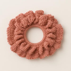 Könnte beinhalten: Ein staubrosafarbenes Scrunchie mit strukturiertem, gestricktem Aussehen. Das Haaraccessoire hat ein gerüschtes Design um das Gummiband. Das Scrunchie ist kreisförmig und scheint aus einem weichen, garnartigen Material zu bestehen.