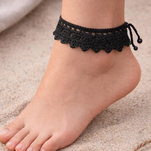 Puede incluir: Una tobillera negra de ganchillo con borde festoneado y cierre de lazo. La tobillera se lleva en un pie que descansa sobre arena. El diseño presenta un delicado patrón calado y los extremos terminan con pequeñas cuentas negras.
