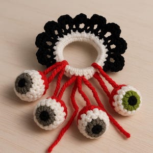Könnte beinhalten: Handgefertigtes Härchen-Accessoire aus Häkelarbeit. Es zeigt einen weiß-schwarzen, gewellten Ring mit hängenden gehäkelten Augäpfeln. Die Augäpfel sind weiß mit grauen oder grünen Iris und schwarzen Pupillen, verbunden durch roten Faden.