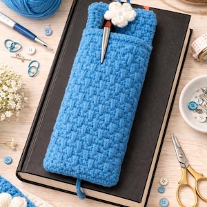 Könnte beinhalten: Eine handgefertigte, hellblaue Häkel-Buchhülle mit einem weißen Blumenakzent und einem Stift in der Tasche. Die Hülle liegt auf einem schwarzen Buch, umgeben von Bastelmaterialien wie Garn, Knöpfen und Scheren.