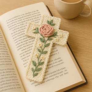 Puede incluir: Marcapáginas de cruz de ganchillo color crema con una rosa rosa y hojas verdes. El marcapáginas está sobre un libro abierto, junto a una taza de café. Un artículo decorativo hecho a mano.