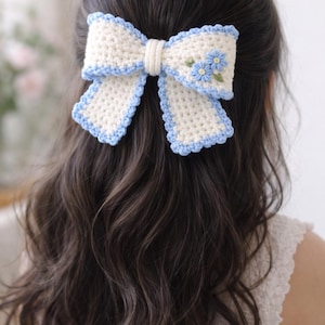 以下が含まれることがあります： クリーム色の手編みヘアリボン、水色のトリムと小さな刺繍の青い花が特徴です。 この装飾的なヘアアクセサリーは、どんなヘアスタイルにも魅力を添えるのに最適です。