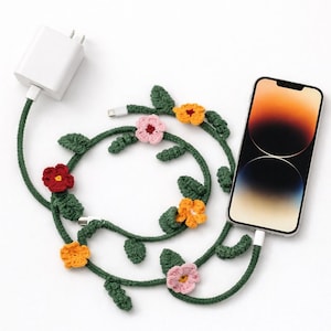 Könnte beinhalten: Ein grünes Häkel-Ladekabel mit floralem Design. Das Kabel ist mit gehäkelten Blättern und bunten Blumen in Rot-, Rosa- und Orangetönen verziert. Ein weißes Ladegerät und ein Smartphone sind mit dem Kabel verbunden.