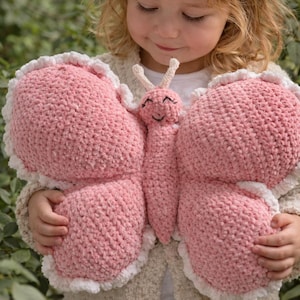Könnte beinhalten: Ein rosa Schmetterlingsspielzeug mit weißen Rändern. Der Schmetterling hat ein lächelndes Gesicht und Fühler. Die Flügel sind groß und abgerundet. Das Spielzeug besteht aus einem strukturierten Material.