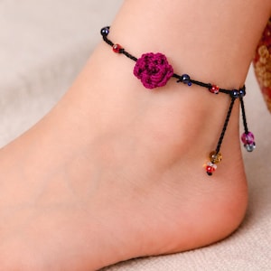 May include: A black anklet featuring a magenta crocheted flower and colourful beads. The anklet is decorated with small red, blue, and yellow beads, and hangs delicately on the ankle. A charming piece of foot jewellery.