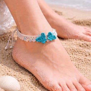 May include: A crocheted ankle bracelet with two turquoise butterfly accents. The bracelet is cream-colored with pastel floral details and a drawstring closure. The image shows the bracelet on a foot on a sandy beach.