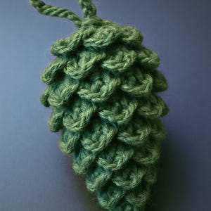Könnte beinhalten: Handgefertigter grüner Häkel-Kiefernzapfen-Ornament mit einer Schlaufe zum Aufhängen. Der Kiefernzapfen besteht aus strukturiertem Garn, und die Schuppen sind deutlich definiert. Eine einzigartige, von der Natur inspirierte Dekoration für die Feiertage oder das ganze Jahr.
