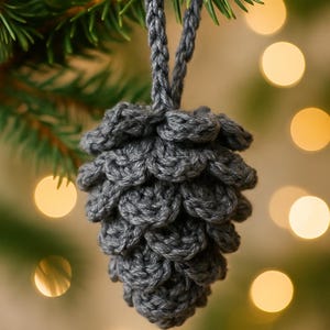 May include: A handcrafted gray crochet pinecone ornament hanging from a Christmas tree branch. The pinecone is made of textured yarn and has a loop for hanging. The background features blurred Christmas lights.