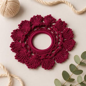 Puede incluir: Una goma para el pelo de ganchillo burdeos con un diseño circular. El accesorio para el cabello presenta un anillo central rodeado por un patrón detallado en forma de flor. El fondo incluye una madeja de lana, una cuerda y vegetación.
