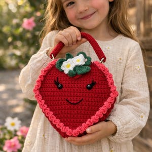 Könnte beinhalten: Eine rote, gehäkelte Erdbeer-Handtasche mit einem lächelnden Gesicht, schwarzen Augen und einem grünen und weißen Blumendetail. Die Tasche hat einen roten Griff und einen rosa gewellten Rand. Die Tasche wird von einer Person gehalten.
