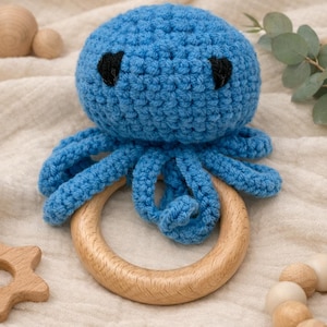 Könnte beinhalten: Ein blauer gehäkelter Oktopus-Babyrassel mit schwarzen Augen und einem Holzring. Der Oktopus hat acht Tentakel und ist für Babys konzipiert. Weitere Holzspielzeuge befinden sich im Hintergrund.