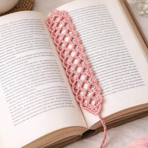 May include: A handmade, light pink crochet bookmark with a diamond pattern, resting in an open book. The bookmark has a tassel at the end. The book is open to a page of text.