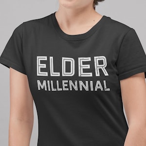 May include: Black t-shirt with the words "ELDER MILLENNIAL" printed in white, block letters. The shirt has a crew neck and short sleeves. The text is centered on the front of the shirt.