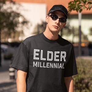 May include: A black short-sleeved t-shirt featuring the white text ELDER MILLENNIAL printed on the chest. The shirt is paired with a black baseball cap and dark-tinted oval sunglasses, creating a coordinated look with a simple, clean aesthetic.