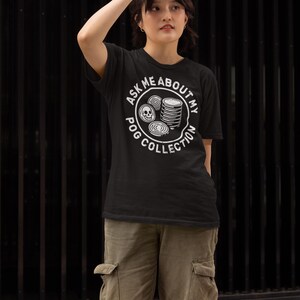 Puede incluir: Camiseta negra con el texto "ASK ME ABOUT MY POG COLLECTION" en un dise&ntilde;o circular blanco. El dise&ntilde;o incluye im&aacute;genes de POGs. La persona lleva pantalones cargo verde oliva.