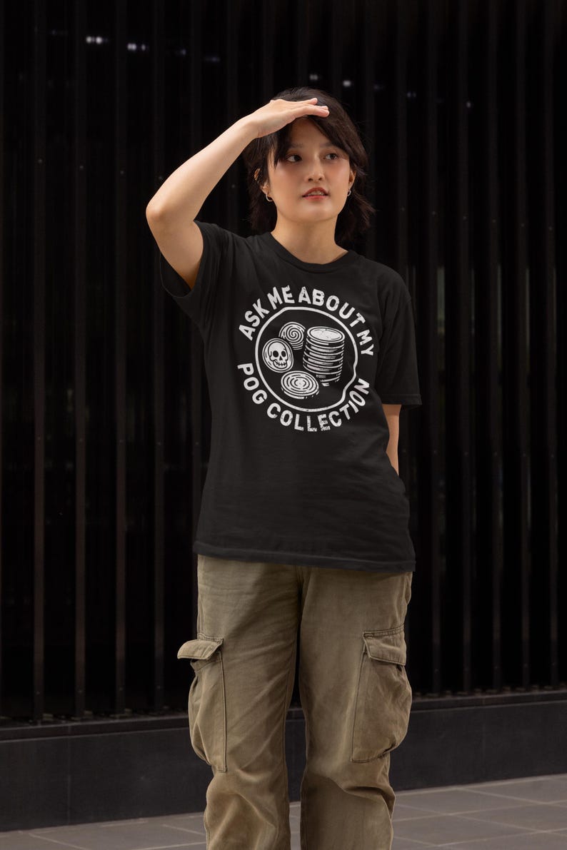 Puede incluir: Camiseta negra con el texto "ASK ME ABOUT MY POG COLLECTION" en un dise&ntilde;o circular blanco. El dise&ntilde;o incluye im&aacute;genes de POGs. La persona lleva pantalones cargo verde oliva.