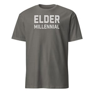 May include: A dark gray short-sleeve t-shirt featuring the text ELDER MILLENNIAL printed in white on the chest. The lettering has a distressed, outlined appearance. The shirt has a crew neckline and a standard fit with a solid color finish.