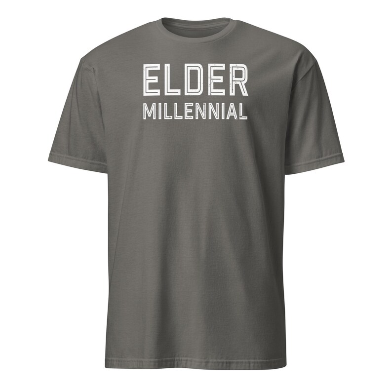 May include: A dark gray short-sleeve t-shirt featuring the text ELDER MILLENNIAL printed in white on the chest. The lettering has a distressed, outlined appearance. The shirt has a crew neckline and a standard fit with a solid color finish.