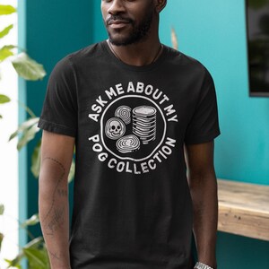 Puede incluir: Camiseta negra con un dise&ntilde;o gr&aacute;fico blanco que dice "ASK ME ABOUT MY POG COLLECTION". El dise&ntilde;o incluye una calavera y pogs apilados. La camiseta es de manga corta.