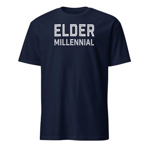 May include: A navy blue short-sleeve t-shirt featuring the text ELDER MILLENNIAL printed in white across the chest. The lettering has a distressed, weathered appearance with a double-line outline style, centered on the front of the garment.