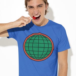 May include: A blue short-sleeved t-shirt featuring a central graphic of a green globe with black grid lines, enclosed within a thick red circular border. The design is printed on the chest of the garment, which has a standard crew neckline.