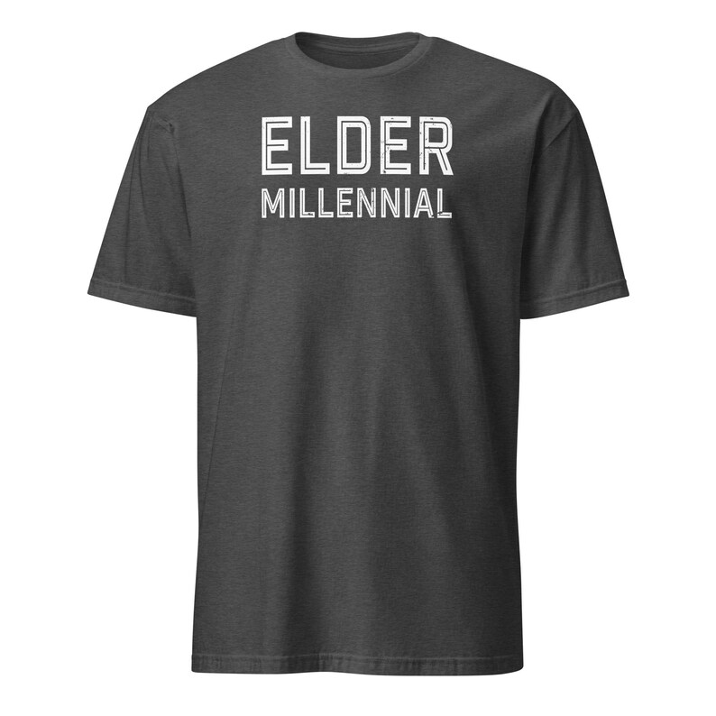 May include: A dark charcoal gray short-sleeve t-shirt featuring the text ELDER MILLENNIAL printed in a white, distressed, sans-serif font across the chest. The garment has a crew neckline and a standard fit with visible fabric texture.