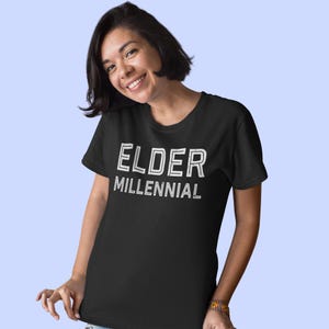 May include: A black short-sleeved t-shirt featuring the text ELDER MILLENNIAL printed in a white, distressed, sans-serif font across the chest. The garment has a classic crew neckline and a straight hemline, providing a simple and casual appearance.