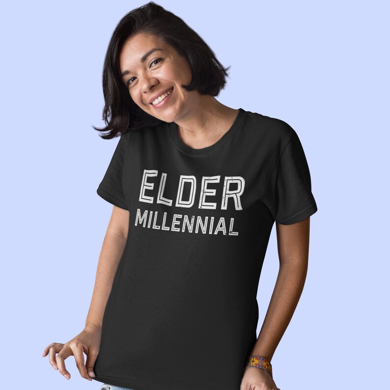 May include: A black short-sleeved t-shirt featuring the text ELDER MILLENNIAL printed in a white, distressed, sans-serif font across the chest. The garment has a classic crew neckline and a straight hemline, providing a simple and casual appearance.