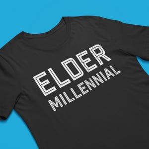 May include: A black short-sleeved t-shirt featuring the text ELDER MILLENNIAL printed in a white, distressed, double-lined font. The text is positioned diagonally across the chest area of the garment, which has a standard crew neckline.