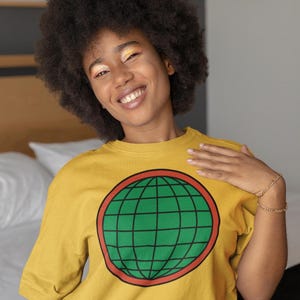May include: A mustard yellow short-sleeved t-shirt featuring a large circular graphic on the chest. The graphic consists of a green globe with black grid lines, enclosed within a thick red circular border. The shirt has a classic crew neckline.
