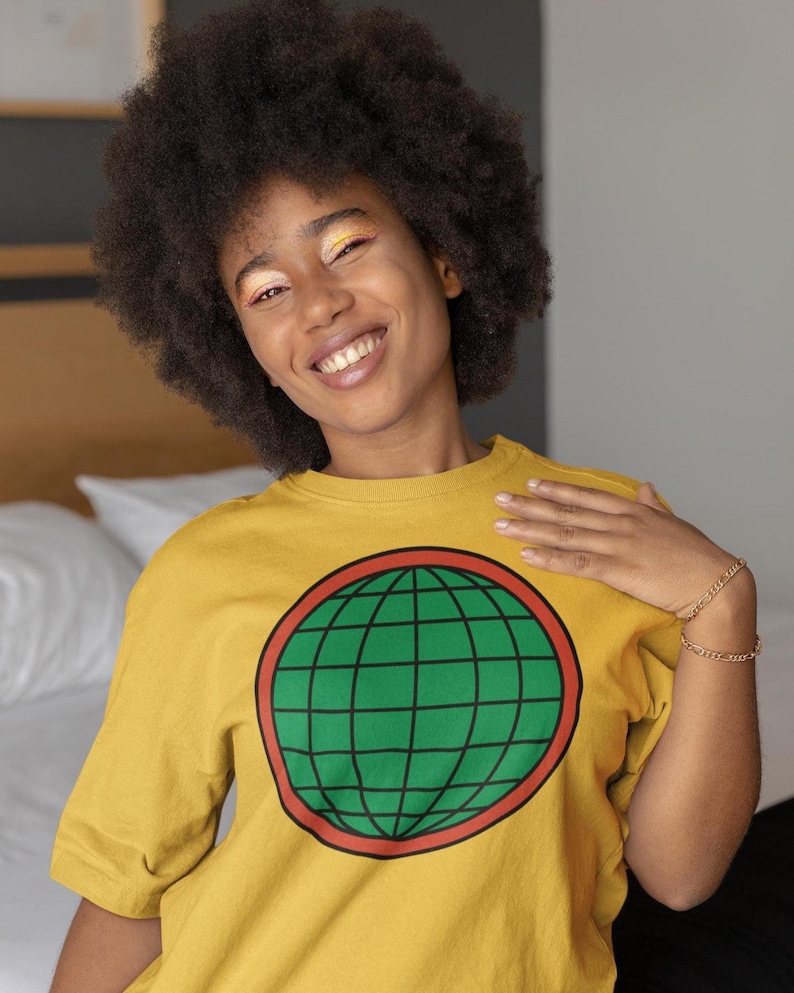May include: A mustard yellow short-sleeved t-shirt featuring a large circular graphic on the chest. The graphic consists of a green globe with black grid lines, enclosed within a thick red circular border. The shirt has a classic crew neckline.