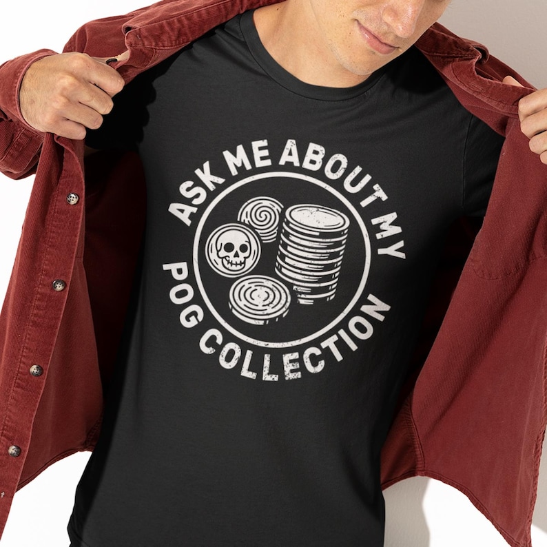 May include: Black t-shirt with a white graphic that says "ASK ME ABOUT MY POG COLLECTION." The graphic includes images of pogs and a skull. The person is wearing a red jacket.