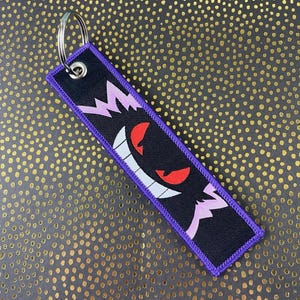 May include: Black and purple embroidered keychain featuring a cartoon ghost with red eyes and a wide grin. The keychain has a silver metal ring and a purple border. The design is on a black background.