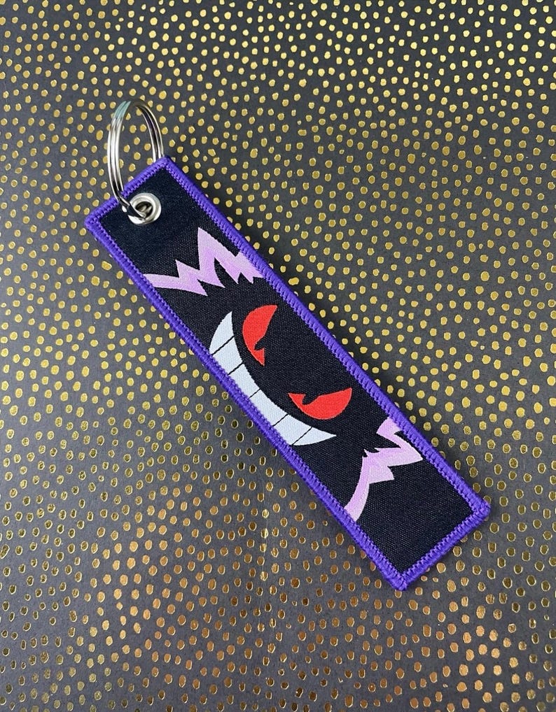 May include: Black and purple embroidered keychain featuring a cartoon ghost with red eyes and a wide grin. The keychain has a silver metal ring and a purple border. The design is on a black background.