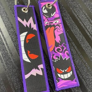 May include: Two black keychains with purple trim and silver key rings. One keychain features a white and red ghost design, and the other has a purple and red monster design. Both keychains have a purple border.