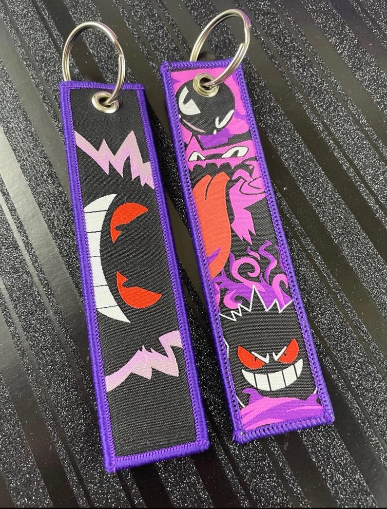 May include: Two black keychains with purple trim and silver key rings. One keychain features a white and red ghost design, and the other has a purple and red monster design. Both keychains have a purple border.