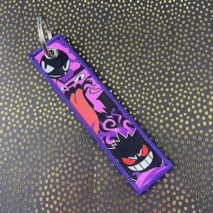May include: A rectangular purple keychain featuring a design of cartoon ghosts in black, pink, and red. The keychain has a silver metal ring for attaching keys. The edges are stitched with purple thread.